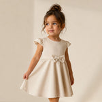 Young girl wearing a beige dress with a bow, standing against a plain background