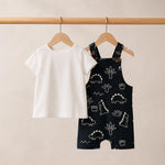 White t-shirt and black overalls with dinosaur pattern on hangers against a beige background