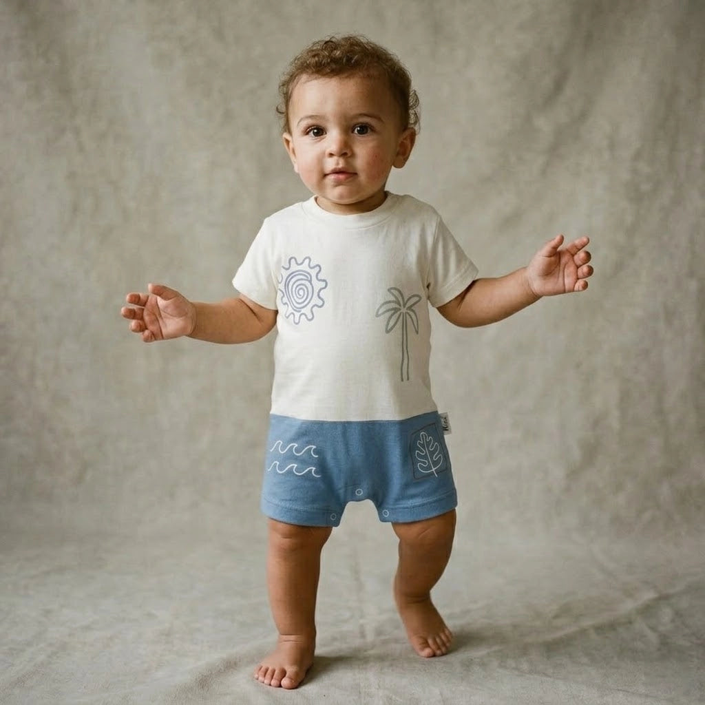 Baby wearing a white shirt with graphics and blue shorts standing next to a beige ottoman on a beige background