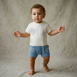 Baby wearing a white shirt with graphics and blue shorts standing next to a beige ottoman on a beige background