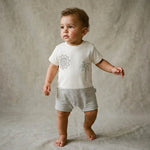 Baby standing next to a beige cube wearing a white t-shirt with graphics and gray shorts on a beige background