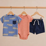 Children's clothing set with a blue shirt, pink onesie, and navy shorts on hangers against a beige background.