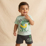 Child wearing a light green t-shirt with a dinosaur and duck design and black shorts with 'Hello Summer' text on a beige background.