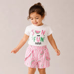 Child wearing a white t-shirt with watermelon design and pink striped shorts on a plain background